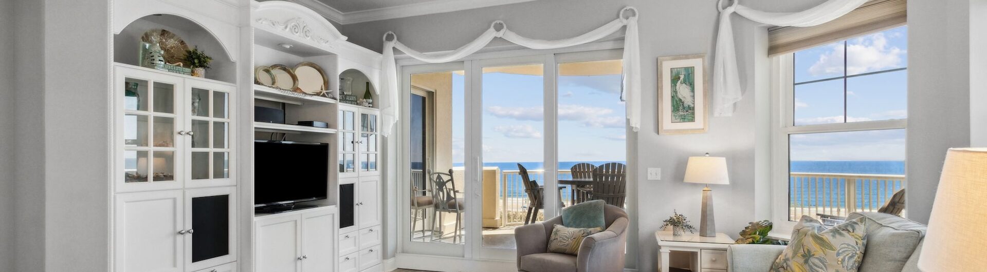 Bright living room with light-colored sofas, a white entertainment center, large windows, and a door opening to a balcony with ocean views. Decor includes coastal accents and patterned pillows.