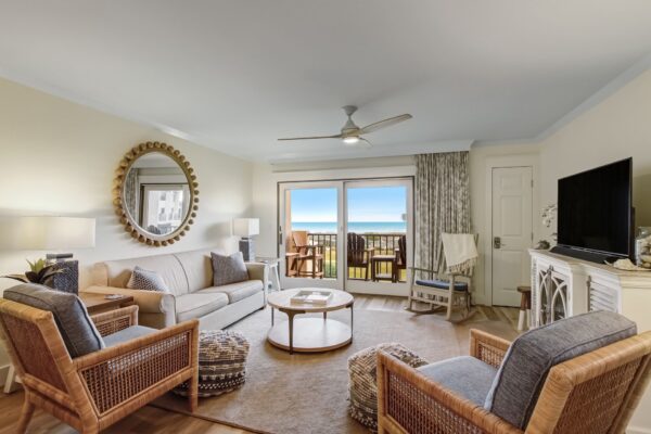 A bright, coastal-themed living room with a sofa, two wicker chairs, a round mirror, a TV, and glass doors leading to a balcony with ocean views. Natural light fills the space, highlighting neutral and blue décor.