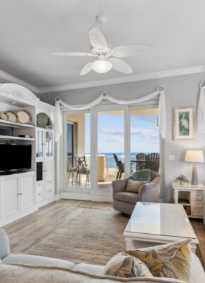 Bright living room with light-colored sofas, a white entertainment center, large windows, and a door opening to a balcony with ocean views. Decor includes coastal accents and patterned pillows.
