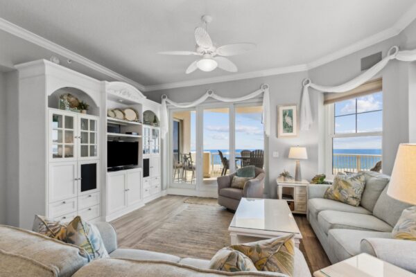 Bright living room with light-colored sofas, a white entertainment center, large windows, and a door opening to a balcony with ocean views. Decor includes coastal accents and patterned pillows.