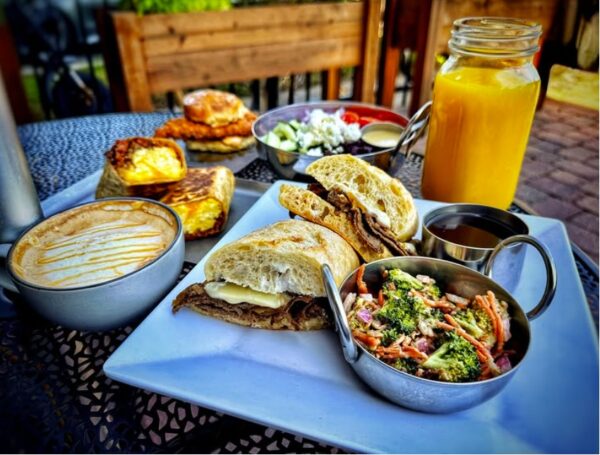 A table with a sandwich, a bowl of broccoli slaw, a cup of coffee, orange juice in a mason jar, scones with jam, and a salad with feta cheese, all served on a metal outdoor patio table.