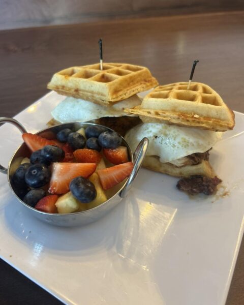 A plated breakfast featuring two waffle sandwiches with eggs and meat, each secured with a toothpick, served alongside a small metal bowl of mixed fresh fruit, including strawberries, blueberries, and bananas.