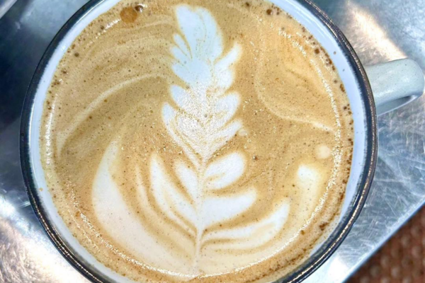 A close-up of a cup of latte with creamy, leaf-shaped latte art on top, served in a white mug on a metal tray.