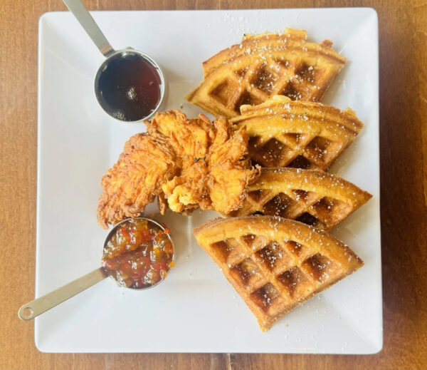 A white square plate with four pieces of golden brown waffle, a fried chicken breast, a small cup of syrup, and a small cup of fruit jam, all arranged neatly on a wooden table.