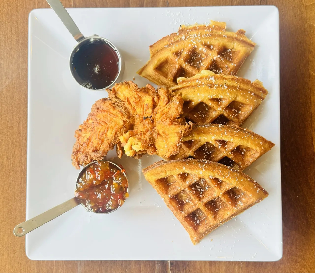 A white square plate with four pieces of golden brown waffle, a fried chicken breast, a small cup of syrup, and a small cup of fruit jam, all arranged neatly on a wooden table.