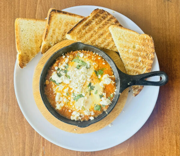 A small skillet of shakshuka topped with crumbled cheese and herbs sits on a wooden board, served with four slices of toasted bread on a white plate, on a wooden table.