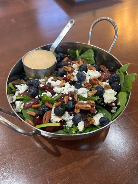 A metal bowl with spinach salad topped with blueberries, pecans, feta cheese, and a small cup of creamy dressing, set on a wooden table.