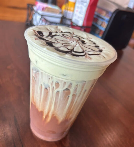 A plastic cup filled with a layered iced drink, topped with creamy foam and chocolate syrup in a decorative swirl pattern. The cup has drips of cream and syrup on the sides, sitting on a wooden table.