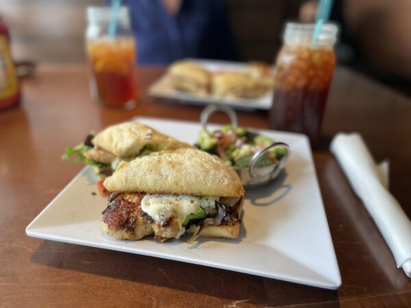 A close-up of a sandwich with grilled chicken, melted cheese, and greens on ciabatta bread, served on a square white plate with a small salad and a drink in the background.
