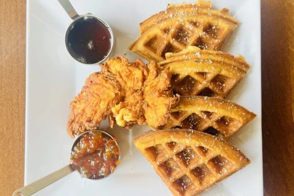 A plate with three waffle quarters, two pieces of fried chicken, a small cup of syrup, and a small cup of fruit preserves, all served on a white dish.