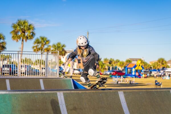 A young child wearing a helmet and protective gear skateboards on a ramp at an outdoor skate park. Palm trees, cars, and people are visible in the sunny background.