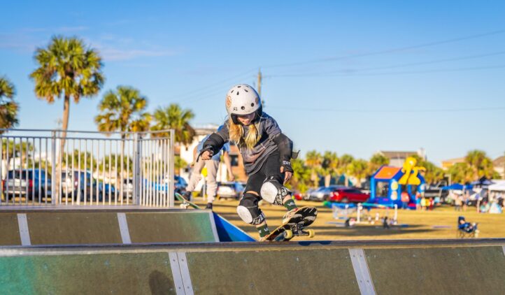 A young child wearing a helmet and protective gear skateboards on a ramp at an outdoor skate park, with palm trees, cars, and people in the background under a clear blue sky.