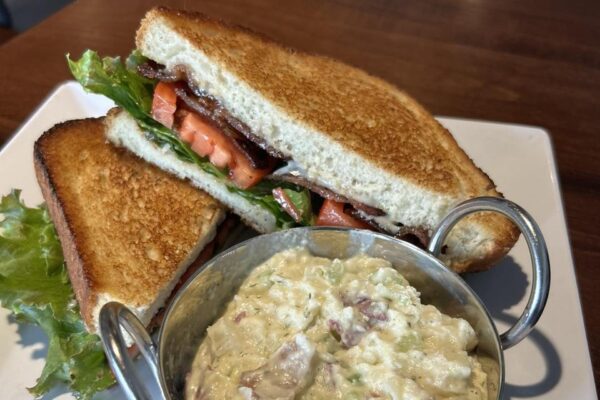 A BLT sandwich with crispy bacon, lettuce, and tomato on toasted bread is served on a white plate with a side of creamy potato salad in a small metal bowl.