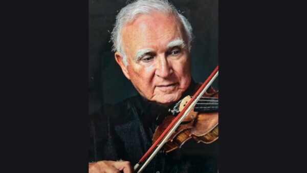 An elderly man with white hair plays a violin, focusing intently on the instrument. He wears a dark shirt and is set against a black background.