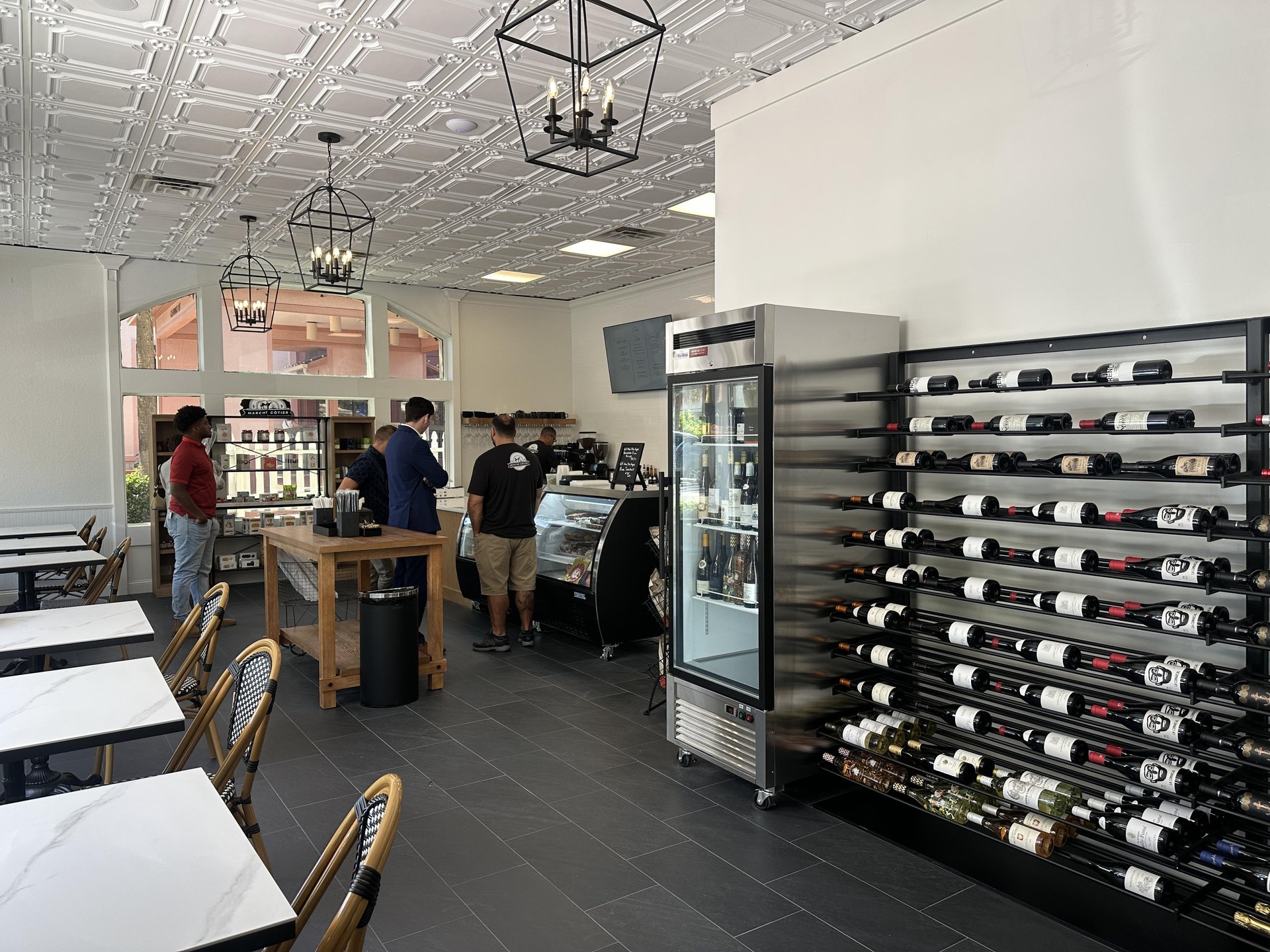 A modern café interior with three people at the counter, wine bottles displayed on racks, a glass refrigerator, white tables with wicker chairs, and decorative white ceiling tiles.