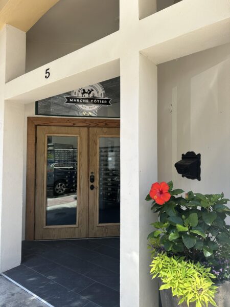 Entrance to a building with a wooden door, large glass panels, and the sign “Marché Cotier” above the doorway. A bright red hibiscus flower and green plants are in a pot to the right of the door. The number 5 is on the wall.