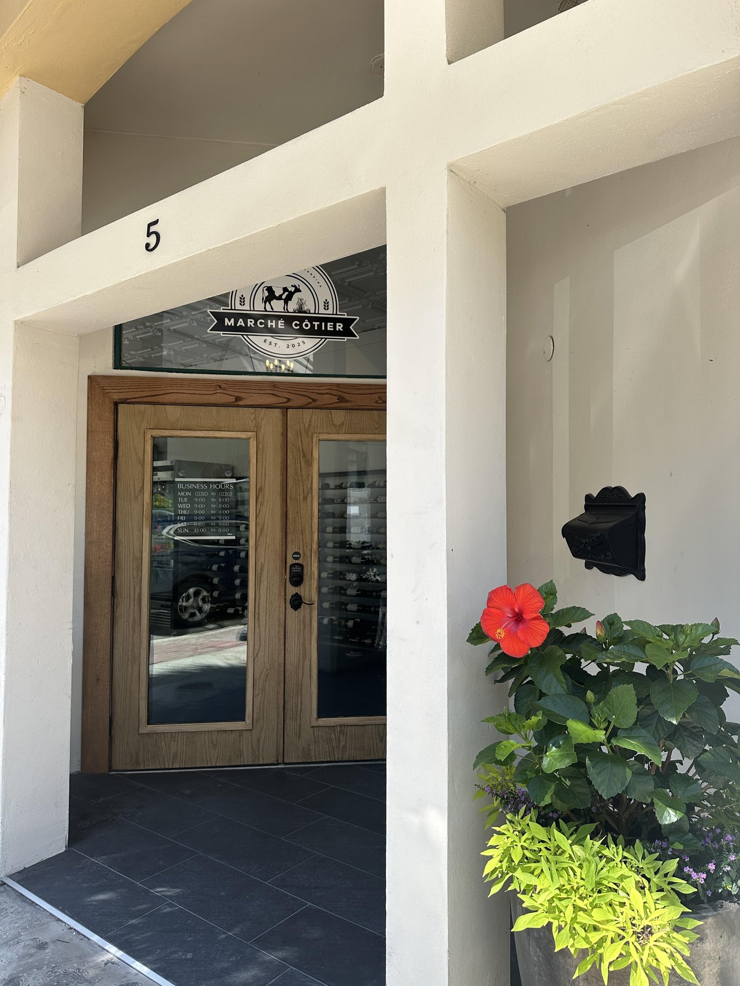 Entrance to a building with a wooden door, large glass panels, and the sign “Marché Cotier” above the doorway. A bright red hibiscus flower and green plants are in a pot to the right of the door. The number 5 is on the wall.