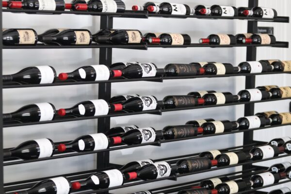 Rows of wine bottles with various labels and red foil tops are neatly arranged on a black metal rack against a white wall. The bottles are stored horizontally in a modern, organized display.