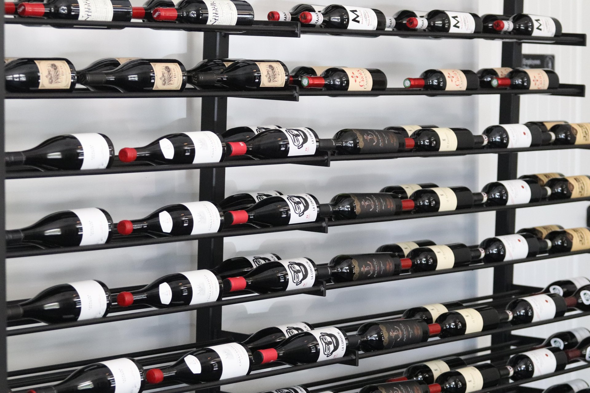 Rows of wine bottles with various labels and red foil tops are neatly arranged on a black metal rack against a white wall. The bottles are stored horizontally in a modern, organized display.