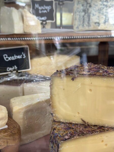 A display of various cheeses behind glass, including a wedge coated with dried herbs or flowers in the foreground and handwritten labels on small chalkboards in the background.