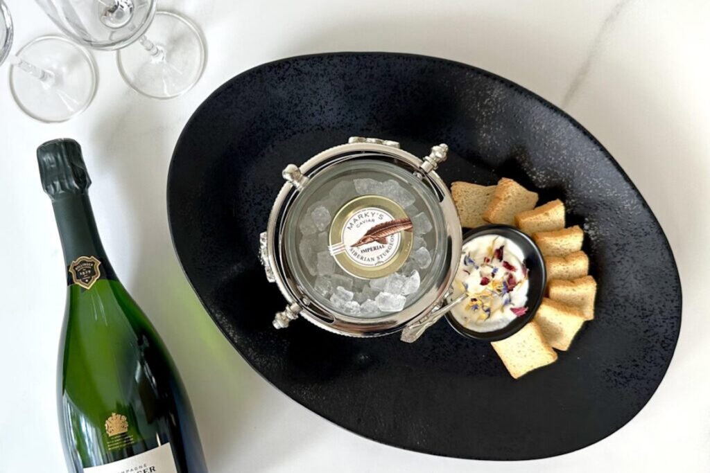 A black oval plate holds a jar of caviar on ice, a bowl of creamy dip with herbs, and crackers arranged in a fan shape. Nearby are two empty wine glasses and a chilled bottle of sparkling wine on a white surface.