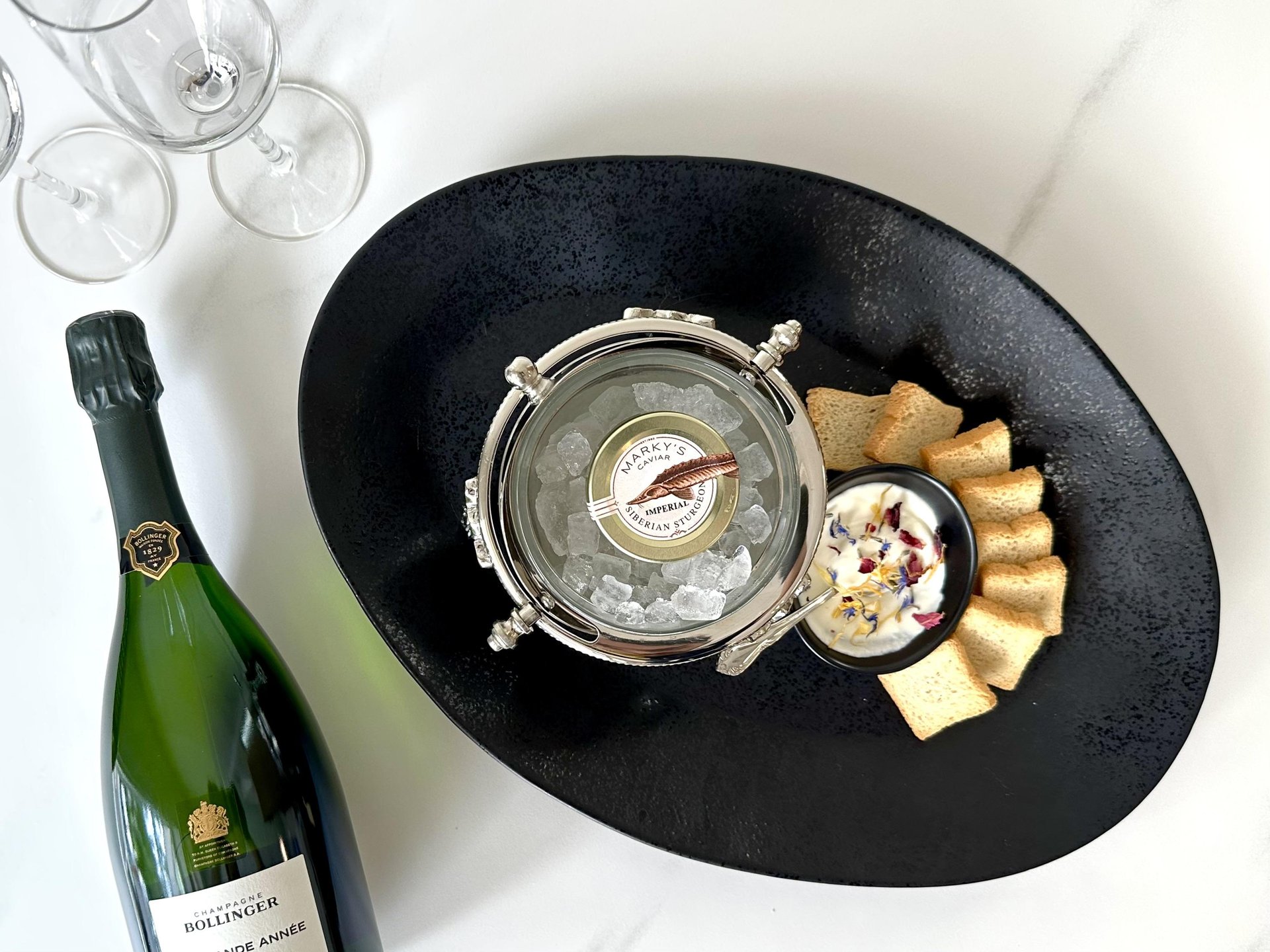 A black oval plate holds a tin of caviar on ice, a small bowl of creamy dip with flower petals, and toast points. Nearby are a bottle of Bollinger champagne and three empty wine glasses on a white surface.