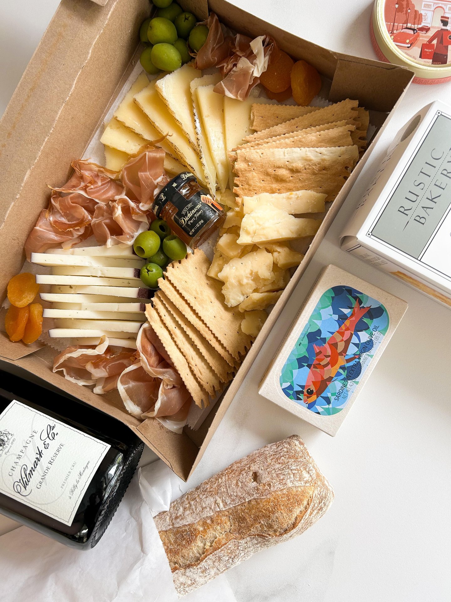A charcuterie box with assorted cheeses, crackers, prosciutto, green olives, dried apricots, fig jam, a rustic bread loaf, a tin with a fish illustration, a box of Rustic Bakery crackers, and a bottle of wine.