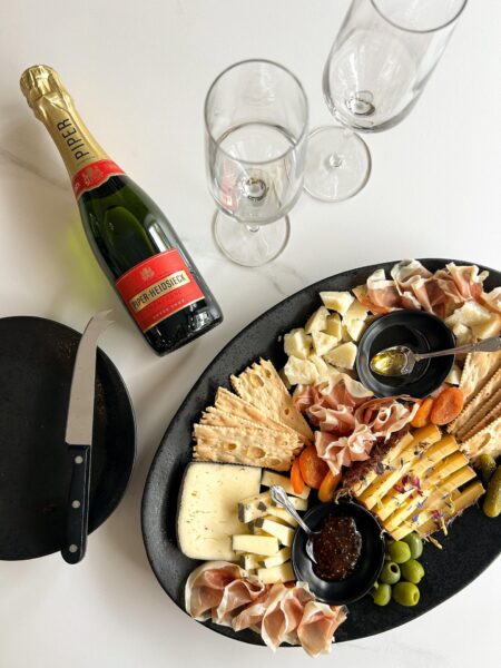 A charcuterie board with assorted cheeses, crackers, prosciutto, olives, dried apricots, and dip sits beside a black plate with a knife, three empty champagne flutes, and a bottle of Piper-Heidsieck champagne.