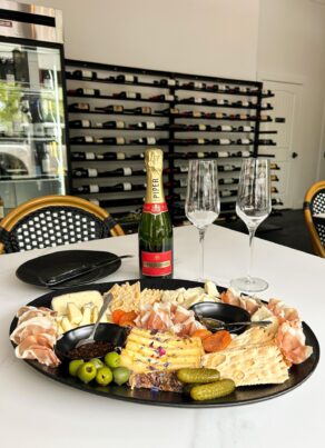 A charcuterie board with assorted cheeses, crackers, meats, olives, and pickles on a table beside a bottle of champagne and two empty flutes, with a wall of wine bottles and café chairs in the background.