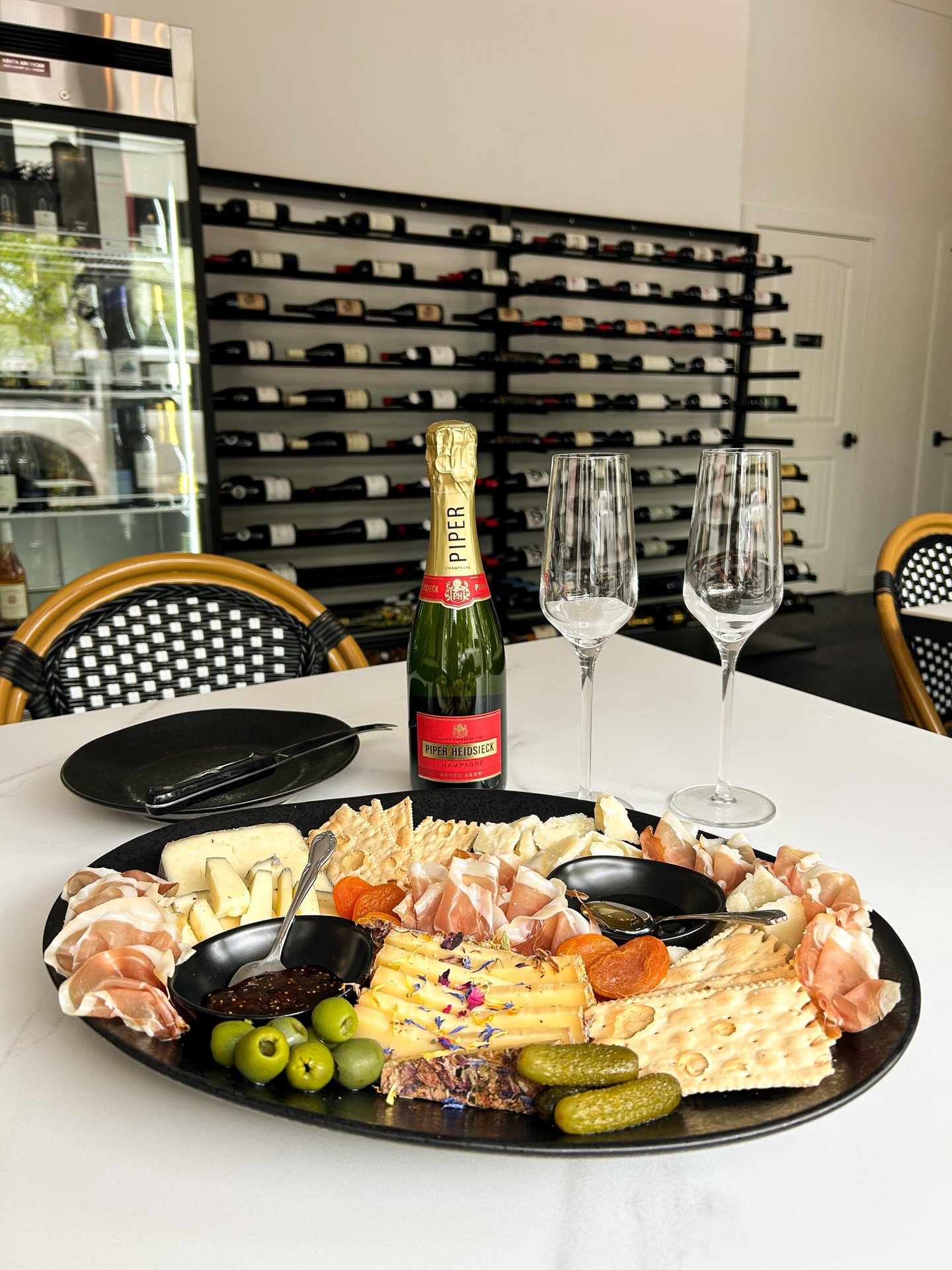 A charcuterie board with assorted cheeses, crackers, meats, olives, and pickles on a table beside a bottle of champagne and two empty flutes, with a wall of wine bottles and café chairs in the background.
