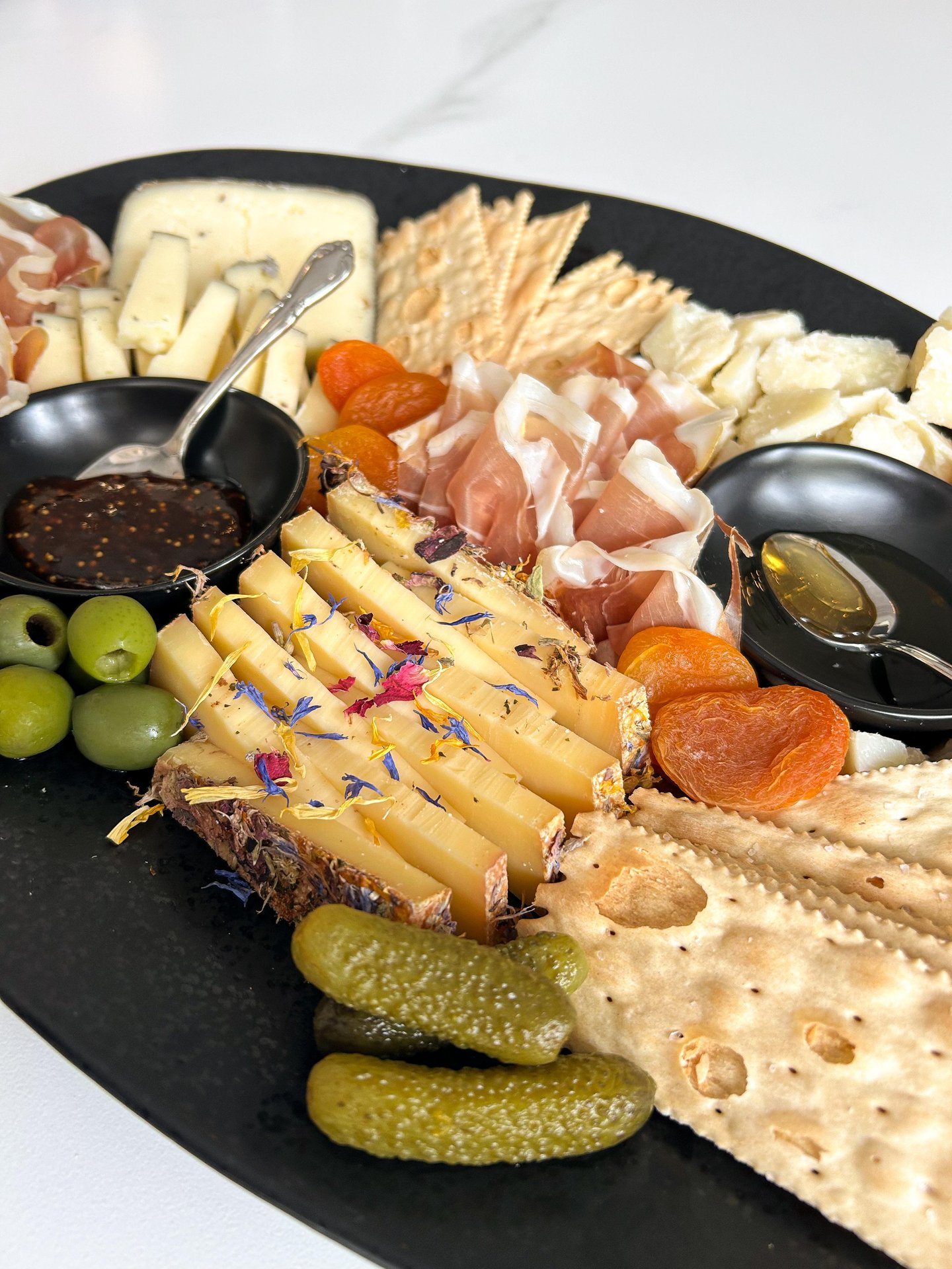 A black platter with assorted cheeses, crackers, prosciutto, green olives, dried apricots, pickles, and two small bowls—one with fig jam, one with honey—presented on a white surface.