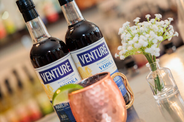 Two bottles of Venture blueberry smash vodka stand on a bar counter next to a copper mug garnished with lime and a small glass vase containing white flowers. The background is softly blurred.