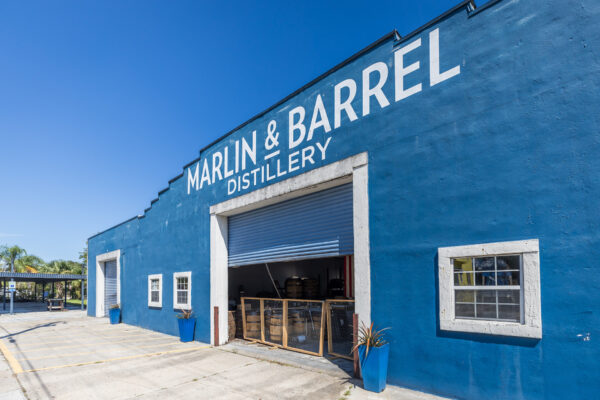 A blue building with white trim and the words Marlin & Barrel Distillery painted on the front, featuring an open garage door revealing barrels inside, under a clear blue sky.