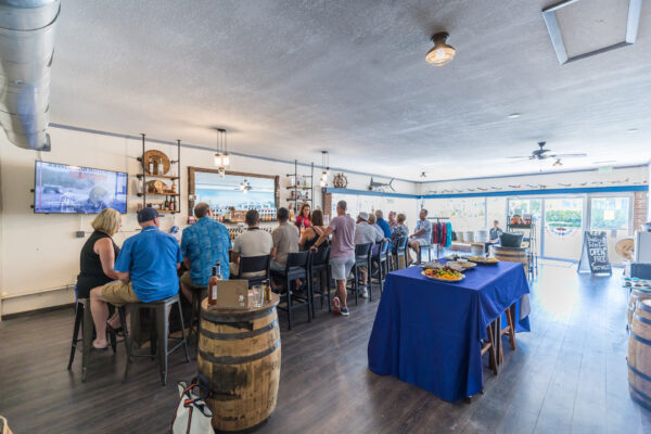 A group of people sit at a bar inside a bright, spacious room with wood floors. Some are eating and drinking. A table with appetizers and a blue tablecloth is in the foreground. Light streams in from large windows in back.