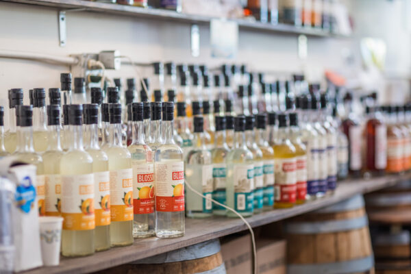 A variety of liquor bottles with colorful labels are neatly arranged on a wooden shelf in a store, with some barrels visible underneath and shelves of additional bottles in the background.