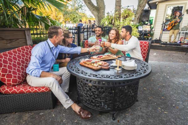 Five people sit around an outdoor table, clinking glasses and smiling. There are platters of food on the table. Trees, a fence, and a musician playing guitar are visible in the background. It appears festive and relaxed.
