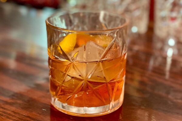 A glass of amber-colored cocktail with large ice cubes and an orange peel garnish sits on a wooden surface. The glass has a textured, geometric pattern.