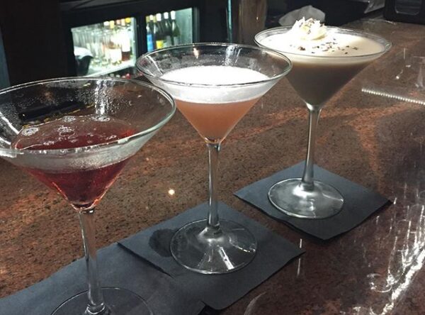 Three different cocktails in martini glasses are lined up on a bar counter, each on a black napkin. The drinks vary in color: one red, one pink, and one creamy white with a garnish on top. A fridge is visible in the background.