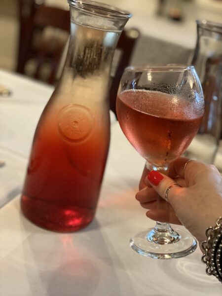A hand with red-painted nails holds a glass of rosé wine next to a matching carafe on a white tablecloth, with blurred chairs and tables in the background.