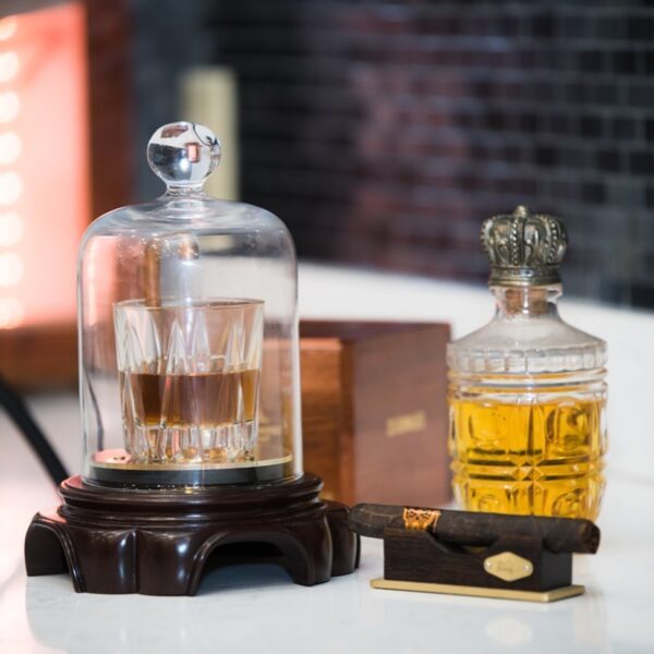 A glass of whiskey under a glass dome sits on a wooden base next to a crystal decanter filled with amber liquid and a cigar on a gold tray, all arranged on a countertop.