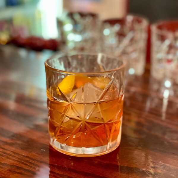 A glass tumbler with a patterned design holds an amber-colored cocktail over large ice cubes and an orange twist, sitting on a polished wooden bar with blurred glasses in the background.