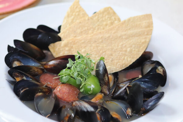 A white plate filled with cooked mussels in broth, garnished with sliced sausage, green herbs, and two tortilla chips standing upright in the center.