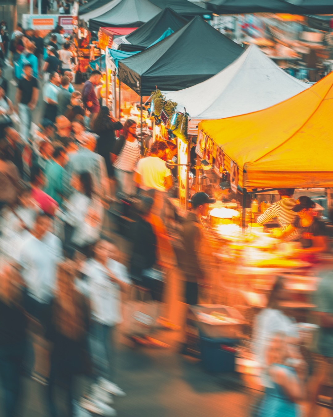 A busy outdoor market scene with colorful tents and blurred crowds of people walking and shopping, creating a lively and vibrant atmosphere.