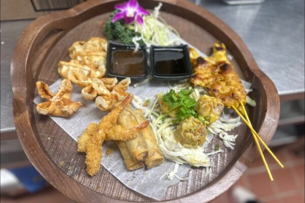 A wooden platter with a variety of Asian appetizers, including fried shrimp, spring rolls, chicken satay skewers, wontons, dumplings, two dipping sauces, shredded cabbage, and a purple flower garnish.