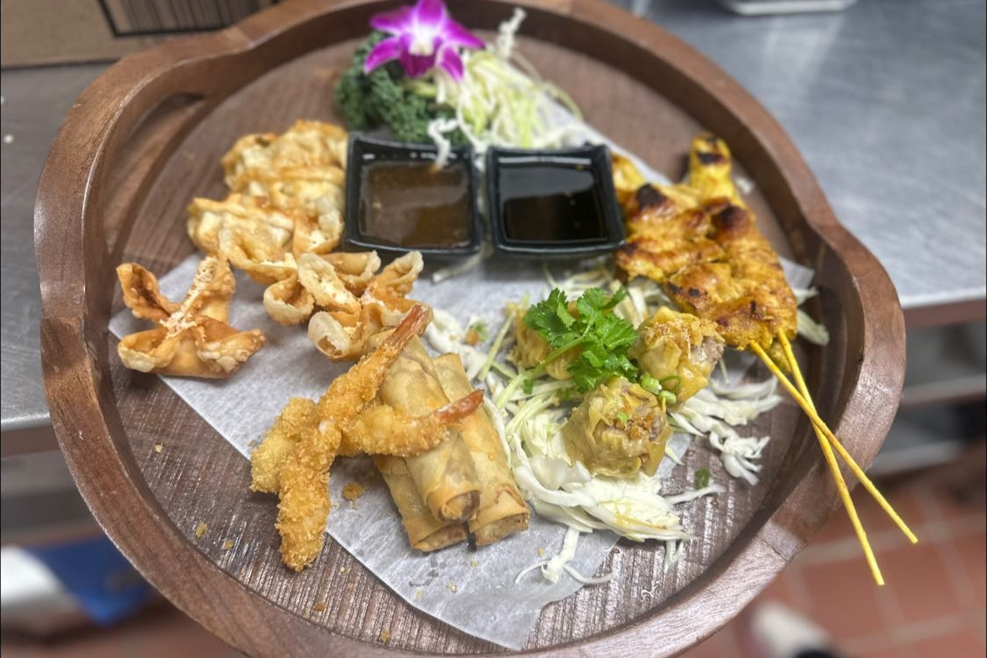 A wooden platter with a variety of Asian appetizers, including fried shrimp, spring rolls, chicken satay skewers, wontons, dumplings, two dipping sauces, shredded cabbage, and a purple flower garnish.
