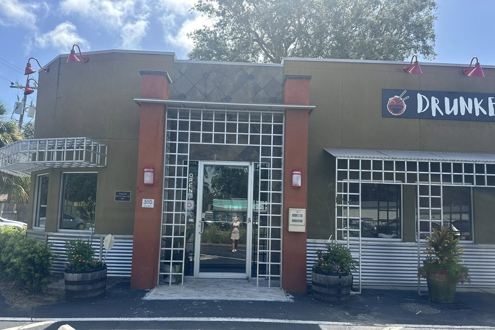 A restaurant exterior with a grid-like metal frame around the entrance, red accents, and two planters. A sign reading “DRUNK” is partially visible. The glass door reflects a person taking the photo.