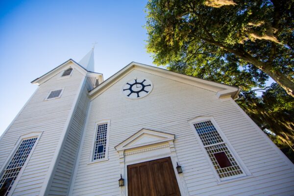 A white wooden church with a tall steeple, large wooden doors, and decorative windows stands under a clear blue sky next to a leafy green tree.