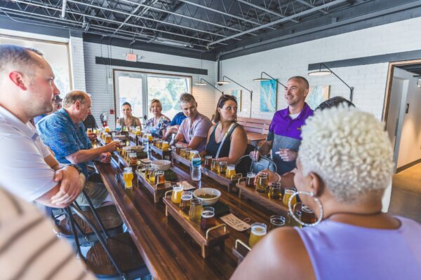 A group of people sit around a long wooden table in a modern brewery, enjoying beer flights and conversation. Natural light streams in through large windows, creating a lively, social atmosphere.