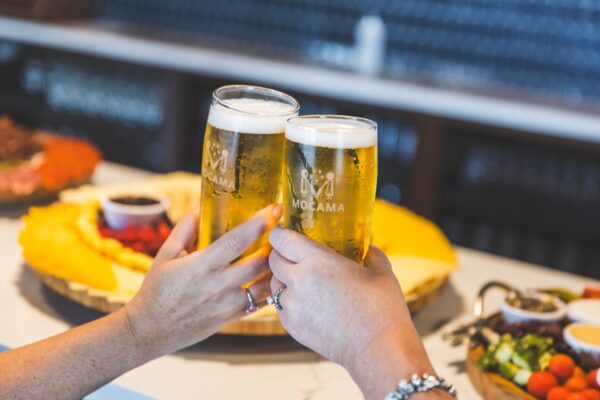 Two people clink glasses of light beer labeled Mocama over a table with assorted food, including a large cheese platter and a plate of fruit and vegetables, in a bright, casual setting.
