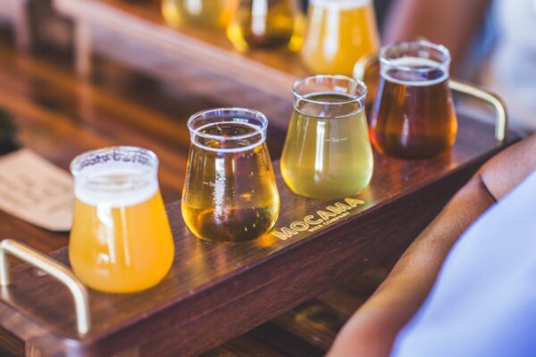 A wooden tray labeled “Mocama” holds four glasses of beer in different shades, ranging from pale yellow to amber, on a wooden table.